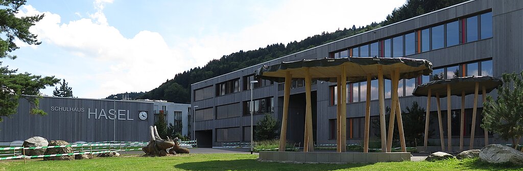 Holzfassade Turnhalle (l., mit Schriftzug), Schulgebäude und Spielplatz.