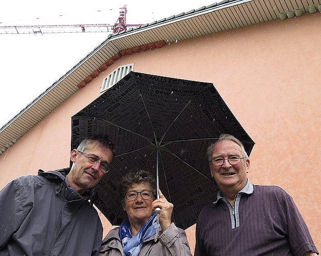 Vogelschützer Andres Beck, Susanne Hobbs, Fernand Herrmann (v.l.). Unter dem Vordach der Turnhalle haben sie Kunstnester für Mehlschwalben (links und rechts, grau) und Mauersegler (im Giebel, rötlich) montiert.Foto: Mü
